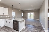 This single-floor unit is designed with an open-concept layout that seamlessly connects the kitchen and living area. The kitchen is well-appointed with crisp white shaker-style cabinetry featuring black hardware, granite countertops, and wood-look vinyl flooring. A peninsula with a double stainless steel sink and built-in dishwasher serves as the central hub, illuminated by two pendant lights with glass cylinder shades. Stainless appliances include a French door refrigerator with ice and water dispenser, an electric range, and an over-the-range microwave. Recessed lighting and a textured ceiling complete the kitchen's functional aesthetic. The adjacent living area features plush carpet and a sliding glass door that opens onto a private balcony with outdoor views. The bedrooms are carpeted, finished with neutral-toned walls, and receive natural light through vertical-blind-covered windows. One bedroom includes a walk-in closet with built-in wood shelving. The bathroom features a tub-shower combination, a granite-topped vanity, and wood-look vinyl flooring, maintaining a consistent material palette throughout the space.