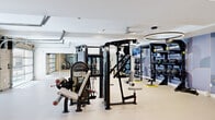 This modern fitness center spans a generous single floor, thoughtfully designed to accommodate a wide range of workout disciplines. The space is organized into distinct functional zones, each serving a specific training purpose. The strength training area features a comprehensive dumbbell rack with a full range of weights, adjustable benches, cable machines, and a dedicated weight plate storage system, all complemented by floor-to-ceiling mirrors that enhance the sense of openness. A separate TRX Training Zone is outfitted with medicine balls, kettlebells, battle ropes, and suspension trainers mounted on a structured storage rack, set against a stylish decorative accent wall. The cardio section houses a variety of machines including stair climbers, ellipticals, and stationary bikes, positioned beneath ceiling fans and ring pendant lighting, with a mounted TV and a wall-mounted mini-split for climate control. A water fountain station is conveniently located nearby. The space features durable light-colored flooring throughout and benefits from abundant natural light, courtesy of two full-panel glass garage doors framed by exposed brick, which open the interior to the outdoors. Recessed lighting and modern circular pendant fixtures maintain a bright, contemporary ambiance throughout.