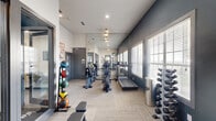 This fitness center is thoughtfully designed to support a comprehensive workout experience within a single-floor layout. The space features a clean, modern aesthetic with bold navy blue accent walls, neutral carpet tile flooring, and large windows fitted with blinds that allow ample natural light to fill the room while offering views of the surrounding outdoors.

The main exercise area is lined with cardio equipment, including multiple treadmills and elliptical machines, arranged along the windowed wall to create an open and airy feel. Full-length mirrors along one wall visually expand the space and allow users to monitor their form during workouts. A wall-mounted television adds to the overall functionality of the cardio zone.

Strength training is well represented with a multi-station cable and weight machine, a flat workout bench, and a tiered dumbbell rack stocked with a range of free weights. A colorful medicine ball rack near the entrance adds versatility for functional fitness routines.

Toward the rear of the space, a built-in cubby storage unit with hooks provides convenient organization for personal belongings. A separate adjoining room, visible through glass partition walls, appears to serve as an office or consultation area. Recessed and ceiling-mounted lighting ensures consistent illumination throughout, completing this practical and well-appointed fitness facility.