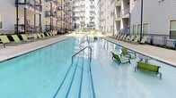 The pool area is a well-appointed outdoor amenity space nestled within a multi-story residential courtyard. The centerpiece is a generously sized swimming pool with clear blue water, framed by clean-lined tile accents and multiple stainless steel entry railings. The expansive concrete pool deck provides ample room for lounging, and is furnished with bright green chaise lounges and accent chairs that add a vibrant contrast to the neutral surroundings. Lush landscaping borders the perimeter of the deck, featuring hydrangeas and various shrubs that soften the hardscape and contribute to an inviting, resort-like atmosphere. The courtyard setting means the pool is enclosed on multiple sides by the building's exterior, offering a sense of privacy and shelter. Upper-level balconies with modern metal railings overlook the pool area, reinforcing the architectural cohesion of the space. A doorway on one side provides direct building access to the pool deck. The open sky above ensures abundant natural light throughout the day, making this pool area a bright and comfortable outdoor retreat.