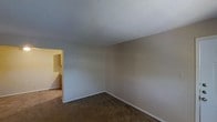 This single-floor space presents a straightforward, functional layout centered around a modestly sized room finished with wall-to-wall carpeting in a warm brown tone. The walls are painted in a neutral beige, creating a clean and understated backdrop that lends itself well to a variety of uses. The textured popcorn ceiling adds a classic touch typical of its construction era. White baseboards frame the perimeter of the room, providing a subtle contrast against the darker flooring. An open doorway on one side connects to an adjacent area, which appears to be lit with warmer tones, suggesting a hallway or secondary room. The overall ambiance is simple and utilitarian, offering a blank canvas ready for personalization. The space is free of ornate detailing, keeping the focus on practical livability. Natural light appears limited based on the visible image, with interior lighting contributing to the overall illumination. The single-floor configuration ensures ease of accessibility and movement throughout the space.