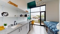 This thoughtfully designed single-floor studio unit makes efficient use of every square foot through smart architectural planning and a clean, contemporary aesthetic. The most striking feature is the oversized floor-to-ceiling window and glass door that floods the interior with natural light and provides direct outdoor access, framing views of surrounding greenery and the streetscape beyond.

A bold teal accent wall adds a vibrant pop of color that anchors the main living area, contrasting beautifully against the crisp white walls and cabinetry throughout. The kitchenette runs along one wall and is outfitted with granite-speckled countertops, white lower cabinets with matte black hardware, an undermount sink with a black faucet, open floating shelves, and a compact under-counter refrigerator. A built-in desk area positioned beside the large window creates a functional workspace bathed in natural light.

The sleeping area features a platform bed with integrated storage drawers, positioned alongside the glass entry door to take advantage of the outdoor views. A modern multi-arm chandelier overhead adds an industrial-modern touch to the space.

Polished concrete floors run throughout the unit, tying together the living, kitchen, and sleeping zones seamlessly. The private bathroom continues the same refined material palette, featuring polished concrete flooring, matte black fixtures, and a clean white toilet, complemented by colorful contemporary wall art.