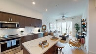 This single-floor residence showcases a thoughtfully designed open-concept layout that seamlessly connects the kitchen, dining, and living areas. The kitchen is a standout feature, outfitted with rich dark-wood cabinetry extending to the ceiling, a full suite of stainless steel appliances including a refrigerator, range, microwave, and dishwasher, and a generous island topped with striking white quartz countertops with grey veining. A classic white subway tile backsplash completes the space. Light wood-tone flooring runs throughout, enhancing the bright, airy atmosphere created by recessed lighting and elevated ceilings.

The living area opens directly onto a private balcony through glass-paneled doors dressed with sheer white curtains, allowing natural light to flood the interior. A ceiling fan adds a casual, coastal touch to this inviting gathering space.

The two bedrooms are comfortably sized, each featuring windows that bring in natural light and views of palm trees and the surrounding landscape. One bedroom includes an accent wall with patterned wallpaper, adding visual depth.

The bathroom is clean and functional, featuring wood-toned cabinetry, a white countertop vanity, light wood-style flooring, and a full tub-shower combination.