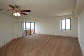 Located at 1 Harbor Ct in Portsmouth, Virginia, this single-floor unit offers a well-organized layout with bright, open living spaces. The expansive main living area is finished with warm wall-to-wall carpeting and features a ceiling fan with light fixture overhead, creating a comfortable and functional environment. Large sliding glass doors open to a balcony, allowing for abundant natural light and panoramic outdoor views of the surrounding cityscape and harbor area. A separate bedroom, also carpeted, includes a wide window framing sweeping elevated views of the city, further enhancing the sense of openness. The kitchen is efficiently designed in an L-shaped configuration and equipped with white cabinetry, granite-style laminate countertops, tile flooring, and a full suite of white appliances including an electric range, over-the-range microwave, dishwasher, and sink. The bathroom is clean and functional, featuring a full soaking tub with a tub/shower combination and chrome fixtures set against white surrounds. Throughout the unit, neutral white walls and consistent finishes create a cohesive, move-in ready aesthetic. The elevated position of the unit provides impressive sightlines and a strong connection to the Portsmouth harbor environment.