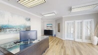 Located at 553 Settlers Landing Rd in Hampton, Virginia, this single-floor commercial space offers a thoughtfully designed layout across a generous footprint. The interior is finished with light hardwood-style flooring, crown molding, and neutral gray walls that create a clean, professional atmosphere throughout.

The main reception and living areas are bright and open, benefiting from abundant natural light streaming through large windows dressed with white blinds and dramatic navy drapes. French doors and glass-panel entryways add an elegant architectural touch, enhancing the sense of openness between zones. Flush, lattice-style ceiling light fixtures provide consistent illumination across the space.

A dedicated dining area features a bold teal accent wall, a glass-top dining table with modern upholstered chairs, and a striking piece of large-format abstract artwork — lending the room a contemporary, curated aesthetic. Adjacent living areas are furnished with comfortable seating arrangements and complementary abstract art, maintaining a cohesive design language.

The kitchen is well-equipped with warm wood cabinetry, granite countertops, and a full suite of stainless steel appliances including a range, microwave, and dishwasher — ideal for an office break room or client-facing hospitality use. A separate office area with a large executive desk and window views rounds out the functional offerings of this versatile Hampton space.