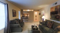 This single-floor residence offers a comfortable and cohesive living environment with a thoughtfully designed open-concept layout. The living room serves as the heart of the space, furnished with plush, dark upholstered sofas arranged around a sleek coffee table, creating an inviting setting for relaxation and entertaining. Warm ambient lighting from a classic table lamp complements the neutral-toned walls, giving the room a cozy and well-appointed feel. Soft wall-to-wall carpeting underfoot adds to the sense of comfort throughout the main living area. The living space flows seamlessly into a raised kitchen bar, which features bar-height seating with stylish wrought-iron stools — ideal for casual dining or conversation. The kitchen, visible beyond the bar counter, is equipped with warm wood cabinetry and standard appliances. A hallway extending from the living area leads to additional rooms, hinting at a practical and well-connected floor plan. Framed artwork on the walls adds a personal touch, enhancing the overall warmth of the interior. The space balances functionality with a welcoming atmosphere, making it a comfortable and livable home.