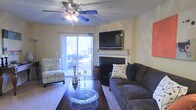 This single-story living space is designed for both comfort and functionality, centered around a clean, neutral color palette with warm beige walls that create an inviting atmosphere. The living room serves as the focal point of the space, anchored by a classic fireplace with a white mantel, above which a wall-mounted flat-screen television is positioned for optimal viewing. A ceiling fan with an integrated light fixture overhead provides both air circulation and ambient lighting throughout the room.

Natural light flows generously into the space through a sliding glass door, which also provides direct access to an outdoor area, offering pleasant exterior views and enhancing the open feel of the room. The flooring features wall-to-wall carpeting, adding warmth underfoot and complementing the cozy aesthetic.

The living area is thoughtfully arranged with a dark upholstered sofa, a patterned accent chair, and a rich wooden coffee table topped with a decorative bowl centerpiece. Tall decorative accents and candle holders add visual interest, contributing to the layered, lived-in character of the space. Overall, the room balances practicality with a welcoming, relaxed ambiance.
