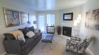 This single-floor space offers a thoughtfully designed living area that balances comfort and style. The living room serves as the focal point of the space, centered around a tile-surround fireplace with a sleek black firebox, accented by a large framed mirror mounted above the mantel. A glass-panel door floods the room with natural light while providing a view of the surrounding greenery outside. The carpeted flooring adds warmth underfoot and complements the neutral, light-toned walls that create a bright and open atmosphere. The living area is generously proportioned, accommodating a full-sized sofa, a tufted ottoman coffee table, and an accent chair with ease. Decorative touches such as framed wall art, a floor lamp, and potted plants contribute to the room's inviting ambiance. A console table positioned near the exterior door provides additional surface space and further enhances the functional layout of the room. The overall design aesthetic is clean and contemporary, with a neutral color palette that lends itself well to a variety of furnishing styles. The combination of natural light, a working fireplace, and a well-considered layout makes this a comfortable and practical living space.