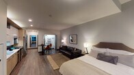 Located at 29 S La Salle St in Chicago's iconic Loop neighborhood, this studio apartment offers a thoughtfully designed open-concept layout that maximizes every inch of its single-floor footprint. The main living area seamlessly integrates sleeping, lounging, and dining functions, featuring dark hardwood-style flooring throughout, recessed lighting, and neutral gray walls that create a calm, modern atmosphere. A full-size bed anchors the sleeping area, complemented by a leather sofa, a woven area rug, and a compact bistro-style dining table with chairs. Wall-mounted artwork adds a contemporary finishing touch. Large windows along one wall frame views of the surrounding Chicago cityscape, allowing natural light to fill the space. The kitchen is a standout feature, equipped with stainless steel appliances including a gas range, dishwasher, and French door refrigerator. Two-tone cabinetry — combining warm wood tones with matte gray uppers — pairs with a striking dark herringbone tile backsplash and a clean white countertop. A dedicated closet area with built-in shelving and hanging rods provides organized storage. The bathroom features a full tub and shower combo with classic white subway tile and light gray floor tile, offering a clean and functional design.
