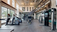 This two-floor fitness facility showcases a striking industrial-chic aesthetic defined by exposed concrete ceilings, visible ductwork, and rubber flooring throughout. The main floor greets visitors with soaring double-height ceilings and floor-to-ceiling windows that flood the space with natural light and offer views of the surrounding greenery. This expansive area houses a comprehensive lineup of cardio equipment, including treadmills, ellipticals, and rowing machines, all arranged to maximize space and flow. A dedicated strength training zone features dumbbell racks and multi-use cable machines, while a functional training area is equipped with wall-mounted rig systems, medicine balls, and TRX suspension trainers set against vibrant teal accent walls. A cubbied storage unit provides convenient organization for personal belongings. A bold geometric wall pattern in teal and natural wood tones adds a distinctive design element throughout. A metal staircase with glass railings connects to the second floor, which opens onto a spacious, light-filled studio with light wood flooring, exposed concrete ceilings, and black-framed glass partitions—ideal for group classes or open training. Together, both floors deliver a well-equipped, thoughtfully designed environment suited to a wide range of fitness activities.