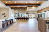 Located in Atlanta, GA (30318), this single-floor community clubhouse showcases a sophisticated blend of modern and warm design elements. The grand entrance welcomes visitors with soaring ceilings clad in natural wood planks, exposed dark steel beams, and large floor-to-ceiling windows and glass doors that flood the interior with abundant natural light and views of surrounding greenery.

The open-concept main area features light-toned large-format tile flooring and flows seamlessly between distinct functional zones. A central island bar, finished in dark cabinetry and topped with a light surface, is lined with cognac-toned swivel barstools beneath a striking geometric patterned accent wall — an ideal social hub. Adjacent, the leasing area presents granite-faced desks paired with stylish leather chairs, set against a richly textured wood accent wall adorned with curated artwork.

A comfortable lounge area offers plush sectional and accent seating, a beverage station, and a sliding barn door leading to additional amenity spaces. A quieter seating alcove provides an intimate retreat with tall windows dressed in linen drapes. For productivity, dedicated work nooks feature built-in floating desks on hardwood floors, accented by an eye-catching gear-motif wallpaper and motivational framed prints. Throughout, thoughtfully selected artwork and designer lighting complete the polished, community-forward atmosphere.