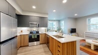 This thoughtfully designed one-bedroom, one-bathroom residence offers a modern open-concept layout across a single floor. The kitchen is a standout feature, showcasing a two-tone cabinetry design with warm wood lower cabinets and sleek matte gray upper cabinets, complemented by white quartz countertops, a classic subway tile backsplash, and a full suite of stainless steel appliances including a range, over-the-range microwave, dishwasher, and French door refrigerator. A generous kitchen island with bar seating serves as both a functional workspace and a casual dining spot. Adjacent to the kitchen, the dedicated dining area benefits from a large window overlooking the surrounding neighborhood, filling the space with natural light. The living room features direct outdoor access through a glass-paned door, along with an additional window that further brightens the space. The bedroom is a comfortable retreat finished with soft carpeting, natural light from a window, and framed wall art, with a dedicated closet nearby for storage. The bathroom continues the home's cohesive aesthetic with warm wood cabinetry, a white vanity countertop, and light wood-look flooring. Recessed lighting and light-toned hardwood-style flooring throughout the main areas tie the space together with a clean, contemporary feel.