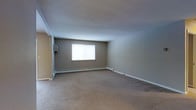 This single-floor space offers a clean and functional layout designed with simplicity and practicality in mind. The main room is generously sized, featuring wall-to-wall carpeting in a warm, neutral tone that adds a sense of comfort underfoot. The walls are finished in a soft greige palette, creating a calm and versatile backdrop suitable for a variety of uses and furnishing styles. A textured popcorn ceiling adds subtle dimension overhead, while white baseboard trim provides a crisp contrast throughout the room. Natural light enters through a centrally placed window, brightening the interior and giving the space an open, airy feel. Baseboard heating units run along the lower walls, ensuring efficient warmth throughout the colder months. The overall ambiance is understated and neutral, offering a blank canvas that can be easily personalized. The space is well-suited for those seeking a straightforward, move-in-ready environment with a practical design and a comfortable, unpretentious character.