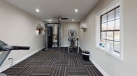 This single-level fitness room offers a comprehensive workout environment designed to accommodate a range of exercise needs. The space is finished with neutral, warm-gray walls and distinctive patterned carpet tiles in a striped, multi-tonal design that adds visual texture underfoot while providing a durable, functional surface suitable for exercise equipment.

The room is outfitted with professional-grade fitness equipment, including a Matrix dual-cable functional trainer with integrated attachments, two treadmills with digital display consoles, and an elliptical cross-trainer. Together, these machines support both cardiovascular and strength training routines.

Overhead, recessed lighting provides bright, even illumination throughout the space, complemented by a ceiling fan that promotes air circulation during workouts. A wall-mounted mini-split climate control unit ensures consistent temperature regulation year-round. Natural light enters through multiple windows, including a large multi-pane window and a glazed entry door, contributing to an open, welcoming atmosphere. The entry features a handsome dark-wood door, adding a warm architectural detail. An informational board is posted near the entrance for user reference.
