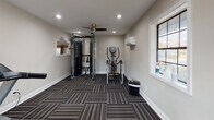This single-level fitness room offers a comprehensive workout environment designed to accommodate a range of exercise needs. The space is finished with neutral, warm-gray walls and distinctive patterned carpet tiles in a striped, multi-tonal design that adds visual texture underfoot while providing a durable, functional surface suitable for exercise equipment.

The room is outfitted with professional-grade fitness equipment, including a Matrix dual-cable functional trainer with integrated attachments, two treadmills with digital display consoles, and an elliptical cross-trainer. Together, these machines support both cardiovascular and strength training routines.

Overhead, recessed lighting provides bright, even illumination throughout the space, complemented by a ceiling fan that promotes air circulation during workouts. A wall-mounted mini-split climate control unit ensures consistent temperature regulation year-round. Natural light enters through multiple windows, including a large multi-pane window and a glazed entry door, contributing to an open, welcoming atmosphere. The entry features a handsome dark-wood door, adding a warm architectural detail. An informational board is posted near the entrance for user reference.