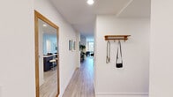 This thoughtfully designed single-floor residence offers five bedrooms, one living room, a contemporary kitchen, and multiple hallways connecting the layout seamlessly. Light wood-tone flooring runs throughout, complementing the crisp white walls and creating a cohesive, modern aesthetic.

The entry hallway sets the tone with a warm wood-framed mirror, a wall-mounted coat rack, and a clear sightline through to the living area. The kitchen is a standout feature, equipped with sleek two-tone cabinetry in dark charcoal and navy, stainless steel appliances, under-cabinet lighting, and a white-topped island with bar seating — ideal for casual dining or meal prep.

The living room is bathed in natural light from floor-to-ceiling windows that frame outdoor views of surrounding greenery and neighboring buildings. The space accommodates comfortable seating and wall-mounted entertainment, with an airy, open atmosphere.

The bedrooms are generously proportioned, each benefiting from large windows — several featuring dramatic floor-to-ceiling glass panels with views of trees and outdoor surroundings. The primary bedroom includes a well-organized built-in closet with shelving and hanging storage. Additional bedrooms offer similar natural light and outdoor views, making each room feel bright and connected to the exterior environment.
