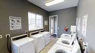 This single-floor shared laundry facility is designed with functionality and cleanliness in mind. The room is finished with cool grey walls and light tile flooring, creating a clean, modern aesthetic. Multiple top-loading washing machines and dryers are arranged along the walls, providing ample capacity for residents. A large window on one wall allows natural light to fill the space while offering a view of the surrounding exterior. A secondary glass-panel door provides an additional point of entry and further enhances the brightness of the room. Safety is addressed with a prominently mounted fire extinguisher on the wall. An informational board is posted near the window, likely displaying usage guidelines or facility rules. The layout is open and efficiently organized, allowing easy access to all machines. The overall ambiance is practical and well-maintained, reflecting a straightforward, utilitarian design intended to serve a shared residential community.