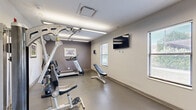 This fitness room at 7820 Lexington Ave in Cleveland, Ohio offers a practical and well-organized exercise space designed to support a full workout routine. The room is finished with neutral gray and white walls, durable rubber-tiled flooring, and bright overhead fluorescent lighting that ensures clear visibility throughout. Natural light filters in through multiple windows fitted with horizontal blinds, adding an open and airy feel to the space.

The room is equipped with a diverse selection of fitness equipment, including a Spirit treadmill, a stationary bike, and a multi-function cable weight machine suitable for a wide range of strength training exercises. A set of dumbbells is neatly racked along one wall, accompanied by a flat/incline adjustable workout bench for versatile strength sessions. A wall-mounted flat-screen television provides entertainment during workouts.

The space is accessed through a glass-paneled door, contributing to a transparent and welcoming entry. Sports-themed framed artwork, including a nod to League Park — a historic Cleveland baseball landmark — adds local character to the room's décor. A security camera is also present, ensuring a safe environment. Overall, this fitness room delivers a compact yet comprehensive exercise facility with a clean, functional aesthetic.
