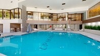 Located at 15 Farm Springs Road in Farmington, Connecticut, this impressive indoor aquatic and leisure facility offers a generously proportioned, single-level layout with a striking double-height mezzanine overlooking the main pool area. The centerpiece is a large, pristine indoor swimming pool with vivid blue water, surrounded by polished concrete decking and an array of modern chaise lounges for relaxed poolside enjoyment. Floor-to-ceiling glass curtain walls and a dramatic skylight ceiling flood the space with abundant natural light, framing views of lush greenery and the outdoor surroundings beyond. A sculptural spiral staircase with sleek metal railings serves as an architectural focal point, connecting the main level to the elevated mezzanine, which features expansive glazed panels that overlook the pool below. Textured accent walls in geometric patterns and warm wood paneling add depth and sophistication to the interior palette. Curated planters of ornamental grasses introduce organic warmth throughout, while recessed and accent lighting enhances the ambiance during evening hours. A dedicated lounge area with white seating and high-top bar tables provides a social gathering space adjacent to the pool. The overall aesthetic blends contemporary design with resort-inspired comfort, creating a refined and welcoming environment suited for both recreation and relaxation.