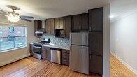 This single-floor space offers a thoughtfully updated interior with a cohesive design throughout. The kitchen serves as a standout feature, showcasing rich dark-stained cabinetry that extends floor to ceiling, offering generous storage. A mosaic tile backsplash in neutral tones adds texture and visual interest, while light granite countertops provide a clean contrast. Stainless steel appliances include a gas range with a wall-mounted range hood, a dishwasher, and a full-size refrigerator, making the kitchen both functional and stylish. Warm hardwood flooring runs throughout the main living areas, lending the space a welcoming and cohesive feel. The living area is bright and open, with multiple windows fitted with blinds that allow for natural light and outdoor views of the surrounding greenery. A ceiling fan adds comfort to the space. The bathroom is well-finished, featuring large white subway tiles in the shower and tub surround, accented by a decorative mosaic border. Wood-look tile flooring and a window with natural light complete the clean, modern aesthetic. Overall, the space presents a practical yet refined environment with updated finishes and a neutral palette throughout.
