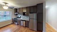 This single-floor space offers a thoughtfully updated interior with a cohesive design throughout. The kitchen serves as a standout feature, showcasing rich dark-stained cabinetry that extends floor to ceiling, offering generous storage. A mosaic tile backsplash in neutral tones adds texture and visual interest, while light granite countertops provide a clean contrast. Stainless steel appliances include a gas range with a wall-mounted range hood, a dishwasher, and a full-size refrigerator, making the kitchen both functional and stylish. Warm hardwood flooring runs throughout the main living areas, lending the space a welcoming and cohesive feel. The living area is bright and open, with multiple windows fitted with blinds that allow for natural light and outdoor views of the surrounding greenery. A ceiling fan adds comfort to the space. The bathroom is well-finished, featuring large white subway tiles in the shower and tub surround, accented by a decorative mosaic border. Wood-look tile flooring and a window with natural light complete the clean, modern aesthetic. Overall, the space presents a practical yet refined environment with updated finishes and a neutral palette throughout.
