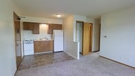 This single-floor space offers a practical, open layout that connects the kitchen and living area seamlessly. The kitchen is equipped with warm medium-toned wood cabinetry featuring simple hardware, a white refrigerator, a white stove, and a single-basin sink with overhead cabinet storage. The kitchen flooring consists of durable tile in a neutral tone, providing a clear visual boundary between the cooking and living areas.

Adjacent to the kitchen, the living space transitions to wall-to-wall carpet, adding warmth underfoot. A white railing separates a small step-down area, adding subtle architectural interest to the layout. Neutral-toned walls throughout lend the space a clean, understated appearance.

Near the hallway, a closet with a rod and shelf provides convenient built-in storage. A wood-toned door leads further into the unit, suggesting additional private spaces beyond the main living area. Flush-mount ceiling lighting provides functional illumination throughout. Overall, the space offers a straightforward, utilitarian design suited for comfortable everyday living.