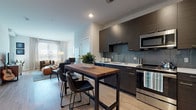 This single-floor residence showcases a thoughtfully designed open-concept layout that seamlessly connects the kitchen, dining, and living areas. The kitchen is outfitted with dark, textured flat-front cabinetry, a subway tile backsplash, light quartz countertops, and a full suite of stainless steel appliances including a range, microwave, dishwasher, and refrigerator. A solid wood-top dining table with an industrial metal base sits adjacent to the kitchen, creating a natural transition into the dining space. The living room is bright and welcoming, with large windows fitted with blinds and sheer curtains that invite abundant natural light. Light-toned hardwood-style flooring flows consistently throughout the entire residence, lending a sense of warmth and continuity. The bedroom is a calm retreat, featuring generous windows that provide excellent natural light and ample space for a full furniture arrangement. The full bathroom is clean and functional, featuring a soaking tub with shower, white tile surround, a dark-cabinetry vanity with a light countertop, and matching hardwood-style flooring. The overall aesthetic is modern and cohesive, balancing dark cabinetry accents with neutral walls and light flooring for a refined, contemporary feel.