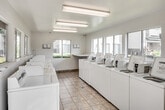 Located at 27550 Hillcrest in Mission Viejo, California, this single-story community laundry facility is designed to serve residents with efficiency and convenience. The space is well-organized, featuring rows of commercial-grade top-load washers and dryers lining both sides of the room, providing ample capacity for multiple users at once. The layout is open and spacious, allowing residents to move comfortably between machines.

Natural light fills the facility through several large sliding windows positioned along the walls, offering views of the surrounding landscaped grounds, mature trees, and the adjacent residential buildings. The bright, airy atmosphere is further enhanced by overhead fluorescent lighting, ensuring the space remains well-lit at all times.

The flooring consists of durable tan ceramic tile throughout, easy to maintain and well-suited for a high-traffic utility space. A built-in countertop with a marble-style surface provides a dedicated area for folding and sorting laundry. Informational notices and usage guidelines are posted on the walls for resident reference. The facility also includes a security camera for added safety. An exterior door offers direct access to the outdoor community grounds, making the space easily accessible and connected to the broader residential environment.