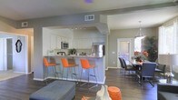 This single-floor residence offers a thoughtfully designed open-concept layout that connects the kitchen, dining, and living areas into one cohesive, flowing space. The kitchen is a standout feature, outfitted with white cabinetry, stainless steel appliances including a refrigerator and microwave, and a generous peninsula with a granite-style countertop. Three vibrant orange bar stools line the peninsula, adding a bold pop of color against the neutral gray walls and dark hardwood-style flooring that runs throughout the space. Track lighting above the kitchen provides focused illumination, complementing the overall well-lit atmosphere. Adjacent to the kitchen, the dining area accommodates a full table and chairs, positioned near a door that invites natural light into the space. The living area, visible in the foreground, features contemporary furnishings that carry through the home's modern aesthetic. Neutral gray tones on the walls create a clean, versatile backdrop, while carefully chosen accent pieces in orange and white add warmth and personality. The open layout enhances the sense of spaciousness, making the home feel inviting and functional throughout.