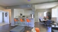 This single-floor residence offers a thoughtfully designed open-concept layout that connects the kitchen, dining, and living areas into one cohesive, flowing space. The kitchen is a standout feature, outfitted with white cabinetry, stainless steel appliances including a refrigerator and microwave, and a generous peninsula with a granite-style countertop. Three vibrant orange bar stools line the peninsula, adding a bold pop of color against the neutral gray walls and dark hardwood-style flooring that runs throughout the space. Track lighting above the kitchen provides focused illumination, complementing the overall well-lit atmosphere. Adjacent to the kitchen, the dining area accommodates a full table and chairs, positioned near a door that invites natural light into the space. The living area, visible in the foreground, features contemporary furnishings that carry through the home's modern aesthetic. Neutral gray tones on the walls create a clean, versatile backdrop, while carefully chosen accent pieces in orange and white add warmth and personality. The open layout enhances the sense of spaciousness, making the home feel inviting and functional throughout.