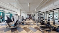 This expansive fitness center is thoughtfully designed to accommodate a full range of workout needs across a single open floor. The space features soaring ceilings with exposed white ductwork and piping, lending an industrial-modern aesthetic that is complemented by bold teal accent walls and a striking geometric-patterned carpet in neutral tones of beige, grey, and charcoal.

The strength training area is generously outfitted with a comprehensive selection of cable machines, weight racks, free weights including a full dumbbell rack, adjustable benches, and multi-functional cable systems. Full-length mirrors line the walls throughout this zone, enhancing both the sense of space and functionality for users monitoring their form.

The cardio section is positioned near a bank of large windows, allowing natural light to fill the area while providing pleasant outdoor views. Modern treadmills and recumbent bikes are arranged to take full advantage of this natural light and scenery.

A dedicated functional training area features stability balls, resistance equipment, and open floor space, defined by deep teal walls and track lighting overhead. A mounted flat-screen television and a water station provide added convenience throughout the facility.

Glass-railed steps connect distinct zones within the space, adding a refined architectural detail. Overall, the fitness center strikes a balance between professional-grade functionality and contemporary design.