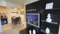 This single-floor residence showcases a thoughtfully designed interior that blends modern aesthetics with functional living. The focal point of the living area is a striking floor-to-ceiling entertainment wall finished in bold black paneling, housing a flat-screen TV and open glass shelves adorned with decorative white accents and candles. The wall unit's dark tone creates a dramatic contrast against the warm, light-colored walls, lending the space a sophisticated, contemporary feel.

The living area flows seamlessly into the dining space, which is visible through an open passageway. The dining area is appointed with a dark wood table and matching high-back chairs, complemented by an elegant chandelier overhead that adds a refined touch. Framed artwork on the walls contributes to the curated, gallery-like atmosphere of the space.

Light hardwood-style flooring runs throughout, unifying the open-concept layout and creating a warm, cohesive foundation. The neutral wall tones allow the darker furnishings and decor elements to stand out, while maintaining an overall sense of brightness and openness. The interplay of dark furniture against light walls and flooring gives the entire space a polished, modern character that is both inviting and visually dynamic.