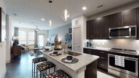 This single-floor residence is thoughtfully designed with a modern aesthetic, combining functionality and style across its open layout. The kitchen serves as a central highlight, showcasing rich dark cabinetry, a subway tile backsplash, granite countertops, stainless steel appliances — including a refrigerator, range, and microwave — and a large island with an undermount sink. Sleek pendant lighting with globe-style fixtures illuminates the island, which doubles as a breakfast bar with seating. The kitchen flows seamlessly into the living area, where polished concrete floors extend throughout and a ceiling fan adds practical comfort. The living room features a soft blue accent wall, large windows with decorative curtains, and abundant natural light. The bedroom is a serene retreat with warm, neutral-toned walls, large windows dressed with patterned curtains and blinds offering exterior views, and ample space for a full furniture arrangement. The bathroom continues the cohesive design language with dark cabinetry, granite countertops, a tiled shower, and clean white walls. Throughout the residence, a consistent palette of dark wood tones, granite surfaces, and light walls creates a polished, contemporary atmosphere.