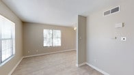 This single-story space is designed with a clean, neutral aesthetic throughout. The interior is unified by light wood-look flooring and warm beige walls, creating a cohesive and understated atmosphere. Textured ceilings add a subtle architectural detail visible in the living and bedroom areas.

The main living area benefits from natural light streaming through a large multi-pane window fitted with blinds, complemented by a ceiling fan with a light fixture overhead. The entry area features a paneled front door alongside an additional window, allowing for cross-ventilation and ample daylight.

A secondary room, likely a bedroom, mirrors the living area's design with its own generously sized window overlooking greenery outside and a dedicated ceiling fan, providing both comfort and views of outdoor foliage.

The kitchen is outfitted with dark espresso-toned cabinetry, light-toned countertops, a double stainless steel sink, and white appliances, offering a practical contrast in finishes. The layout provides adequate counter and storage space.

The bathroom includes a full tub and shower combination, a toilet, a vanity with a speckled countertop, and a large mirror, all finished in clean white tones for a bright, functional feel.