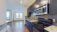 This single-floor residence offers a thoughtfully designed layout that balances style and functionality. Rich hardwood-style flooring flows seamlessly throughout the entire space, creating a warm and cohesive aesthetic.

The kitchen is a standout feature, equipped with dark espresso cabinetry, granite-style countertops, and a suite of stainless steel appliances including a microwave, electric range, and refrigerator. A mosaic tile backsplash adds a refined decorative touch, while ample upper and lower cabinetry provides generous storage.

The kitchen opens directly into a bright and airy living area, where multiple large windows allow abundant natural light to fill the room. A ceiling fan overhead adds comfort and circulation. The open-concept flow between the kitchen and living space makes the area feel expansive and inviting.

The bathroom is well-appointed with dark cabinetry, a granite-topped vanity, a large wall mirror, and a glass-enclosed walk-in shower with full-height tile surround. Adjacent to the bedroom area is a spacious walk-in closet outfitted with multi-tier wire shelving, offering excellent organization and storage capacity.

Overall, the space presents a clean, modern interior with consistent finishes and practical design throughout.