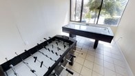 This single-floor game room is designed for recreation and social engagement, offering a well-appointed space for leisure activities. The room is anchored by two prominent game tables — a full-size foosball table and a sleek Playcraft air hockey table — both finished in bold black, lending the space a modern, sporty aesthetic.

Natural light floods the room through large floor-to-ceiling windows that line one wall, providing bright, open illumination throughout the day. The windows also frame picturesque views of mature trees and outdoor greenery, creating a refreshing connection to the outdoors. The light tile flooring complements the clean, minimalist look of the white walls, keeping the atmosphere open and airy.

The layout of the room is thoughtfully arranged to provide ample space around each game table, ensuring comfortable play and movement. The combination of natural light, neutral tones, and purpose-built recreational furnishings makes this game room a functional and inviting destination for players of all ages. Whether enjoying a competitive match of foosball or a fast-paced round of air hockey, this space is well-suited for entertainment and social activity.