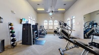 This fitness center offers a thoughtfully designed workout space spread across a single floor, providing ample room for a comprehensive range of exercise equipment. The space is characterized by impressively high ceilings that lend an open, airy atmosphere, enhanced by ceiling fans that ensure comfortable airflow throughout.

Architecturally, the facility features a row of elegant arched windows along the primary wall, allowing abundant natural light to flood the interior while offering views of the outdoors. The flooring throughout is finished in a striking herringbone pattern, adding a refined aesthetic touch to the functional environment.

The main workout area is well-stocked with a diverse selection of cardio equipment, including Matrix treadmills, elliptical machines, a rowing machine, and a stair climber. Strength training needs are addressed by multiple Body-Solid resistance machines, adjustable benches, and a free-weight dumbbell rack. Wall-mounted flat-screen televisions are positioned throughout for entertainment during workouts.

A secondary area toward the rear of the space provides additional strength training stations, along with access to restroom facilities. Recessed and overhead lighting supplements the natural light, ensuring the space remains well-lit at all hours. The overall ambiance is clean, bright, and professional, making it a fully functional and inviting fitness destination.