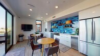 Located at 2048–2068 Carleton St in Berkeley, CA, this modern communal amenity space combines a well-appointed kitchenette and social lounge with seamless access to an expansive outdoor rooftop terrace. The interior is anchored by a striking kitchenette wall clad in vivid blue geometric mosaic tiles that serve as a bold focal point. White flat-front cabinetry, a stainless steel farmhouse sink, a dishwasher, a built-in oven, and warm wood open shelving complete the functional yet design-forward kitchen area. Light hardwood-style flooring runs throughout, complemented by patterned area rugs that define the casual dining zones. Two round wooden tables surrounded by upholstered chairs provide comfortable seating for small groups, while a wall-mounted flat-screen TV with a soundbar adds an entertainment option. Recessed lighting illuminates the clean white walls, and large black-framed windows bring in natural light. Floor-to-ceiling sliding glass doors open directly onto a spacious rooftop terrace finished with large-format porcelain pavers. The terrace is equipped with a gas grill, bar-height dining, lounge seating around a fire pit, and string lights overhead — all framed by sweeping views of the Berkeley hills and the Bay Area skyline beyond.