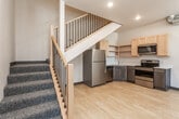 This two-story space offers a functional and modern layout spread across two well-defined levels. The main floor centers around an open kitchen tucked beneath the staircase, efficiently utilizing the available footprint. The kitchen is equipped with a full suite of stainless steel appliances, including a refrigerator, electric range, built-in microwave, and dishwasher. Light maple upper cabinetry is paired with gray lower cabinets and clean white countertops, while open shelving adds an additional storage option. Light hardwood flooring runs throughout both levels, contributing to a cohesive and bright aesthetic.

The staircase, featuring a wood handrail and black metal balusters with blue carpet treads, connects the floors and serves as a visual focal point. The upper level houses the sleeping area, which is generously equipped with two side-by-side sliding-door closets with built-in rods and shelving, providing ample storage. A tall, narrow window in one room allows natural light and outdoor views of the surrounding streetscape. The bathroom is clean and contemporary, featuring a dark wood vanity with a white countertop, a glass-enclosed shower, and light hardwood flooring. Exposed silver ductwork adds an industrial design element throughout.