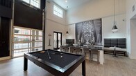 The Baxter's amenity space is a thoughtfully designed, single-floor social hub that combines contemporary style with inviting comfort. Soaring double-height ceilings define the main gathering area, flooding the space with natural light through clerestory windows positioned high along the walls. Large black-framed garage-style doors open fully to an outdoor terrace, seamlessly blending indoor and outdoor living.

At the center of the room, a sleek billiards table anchors the entertainment zone, complemented by a long bar-height counter with modern stools — an ideal setting for socializing. A dramatic oversized black-and-white photographic mural of a bridge serves as a striking focal point on the far wall, lending a strong urban character to the space.

Adjacent to the game area, a comfortable lounge zone features plush seating arranged around a fireplace set against a deep teal accent wall, with a mounted television overhead and a modern globe chandelier adding warmth and ambiance.

The entry area transitions smoothly into the space via herringbone-patterned hardwood flooring, a channel-tufted banquette in warm blush tones, and curated wall art against a bold navy accent wall. Throughout, the design palette of dark metals, light wood floors, and monochromatic artwork creates a cohesive, upscale atmosphere ideal for both relaxed gatherings and lively social events.