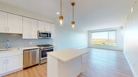 Located along Chief Justice Cushing Highway in Scituate, Massachusetts, this single-floor unit presents a clean, contemporary design throughout. The open-concept layout seamlessly connects the kitchen and living area, creating a bright and cohesive space ideal for both everyday living and entertaining. Light wood-tone flooring runs continuously throughout, lending warmth and visual continuity to the interior.

The kitchen is well-appointed with white shaker-style cabinetry extending to the ceiling, a soft blue-gray subway tile backsplash, and a full suite of stainless steel appliances, including a gas range, built-in microwave, dishwasher, and refrigerator. A central island with a crisp white countertop offers additional workspace, while two brass pendant lights with glass shades add a refined decorative touch overhead.

The living area is generously proportioned, bathed in natural light from a large picture window that frames scenic views of the surrounding landscape. The bedrooms feature the same light hardwood flooring, clean white walls, and ample closet space. The bathroom is finished with a white vanity, integrated sink, tile flooring, and a frameless mirror, maintaining the unit's consistent modern aesthetic throughout.