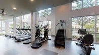 This well-appointed two-story fitness center offers a comprehensive workout experience within a bright, modern facility. The ground floor is anchored by a dramatic double-height cardio zone, where soaring ceilings and large floor-to-ceiling windows flood the space with abundant natural light and provide views of the surrounding landscaped grounds, including palm trees and outdoor amenities. This area is equipped with a full lineup of commercial-grade treadmills, elliptical machines, stair climbers, and stationary bikes, all arranged to maximize both space and sightlines.

A dedicated free weights room on the ground level features an extensive dumbbell rack, weight benches, and wall-to-wall mirrors that visually expand the space, along with a ceiling fan and wall-mounted television for an enhanced workout environment. An additional training alcove houses a multi-function cable trainer and supplemental equipment for functional fitness routines.

The second floor offers its own workout zone, accessible via a central staircase, with open railings that overlook the main floor below. This level includes strength machines, a cable trainer, and additional cardio equipment, along with mounted televisions throughout. A dedicated spin studio with cycling bikes and mirrored walls completes the upper level. Dual water fountains and recessed lighting throughout contribute to the facility's polished, amenity-rich design.