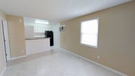 This single-floor junior unit is designed for efficient, comfortable living, featuring a straightforward layout that maximizes its compact footprint. Light beige walls and large-format tile flooring run throughout the space, creating a clean, cohesive aesthetic that reflects natural light effectively.

The open living area connects directly to the kitchen, which is equipped with white shaker-style cabinetry, granite-patterned countertops, a black refrigerator, a black gas range, and a single-basin sink. A peninsula counter separates the kitchen from the living space, adding functionality. A wall-mounted air conditioning unit services the living area, while a fluorescent overhead light provides additional illumination.

The bedroom is a well-proportioned room featuring a ceiling fan with light fixture, a wide window with blinds offering outdoor views and natural light, and the same tile flooring found throughout the unit. A separate door provides access to the exterior.

The bathroom is clean and straightforward, featuring a full tub-shower combination with white tile surround, a toilet, and a vanity with storage. Textured ceilings are present throughout, adding a traditional finish to the overall interior.