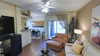 This single-story space features an open-concept layout that seamlessly connects the living area, dining space, and kitchen. The living room is anchored by warm wood-style flooring and a ceiling fan with integrated lighting, contributing to a comfortable, well-ventilated atmosphere. A sliding glass door floods the space with natural light and provides direct access to an outdoor patio, where greenery is visible, enhancing the indoor-outdoor connection.

The living area is generously sized, accommodating a cushioned armchair with matching ottoman and additional seating, arranged around a round coffee table. A tall indoor plant adds a natural, organic element to the corner of the room.

Adjacent to the living space, the dining area is defined by a pendant light fixture overhead and features a dark wood dining table with matching chairs, set atop a low-pile area rug. A decorative colorful artwork on the wall adds visual interest. The kitchen is partially visible beyond a raised counter, suggesting an open, connected layout throughout. Warm, neutral wall tones and thoughtful décor, including a "HOME" wall display, give the space an inviting, lived-in character.