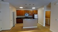 This single-floor space is centered around an open-concept kitchen that seamlessly connects to the adjoining living area. The kitchen is well-appointed with warm medium-toned wood cabinetry that lines the walls, providing generous storage from counter height to ceiling. Dark granite or granite-style countertops offer a striking contrast against the cabinetry, running along the perimeter and continuing onto the central island. The island serves as a functional focal point, ideal for meal preparation or casual dining, and features the same dark countertop surface.

Stainless steel appliances, including a refrigerator, range, and built-in microwave, contribute a modern, polished look to the space. Overhead, a trio of sleek pendant lights hang above the island, complementing the recessed ceiling fixtures that provide ambient illumination throughout the kitchen. The flooring transitions from tile in the kitchen area to neutral carpeting in the adjoining living space, creating a clear yet open delineation between zones. The overall color palette—warm wood, dark counters, and neutral walls—gives the space a balanced, contemporary feel.