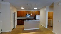 This single-floor space is centered around an open-concept kitchen that seamlessly connects to the adjoining living area. The kitchen is well-appointed with warm medium-toned wood cabinetry that lines the walls, providing generous storage from counter height to ceiling. Dark granite or granite-style countertops offer a striking contrast against the cabinetry, running along the perimeter and continuing onto the central island. The island serves as a functional focal point, ideal for meal preparation or casual dining, and features the same dark countertop surface.

Stainless steel appliances, including a refrigerator, range, and built-in microwave, contribute a modern, polished look to the space. Overhead, a trio of sleek pendant lights hang above the island, complementing the recessed ceiling fixtures that provide ambient illumination throughout the kitchen. The flooring transitions from tile in the kitchen area to neutral carpeting in the adjoining living space, creating a clear yet open delineation between zones. The overall color palette—warm wood, dark counters, and neutral walls—gives the space a balanced, contemporary feel.