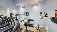 This fitness center offers a practical and well-organized space designed to accommodate a variety of workout routines. The room is finished with soft blue walls and recessed lighting, creating a bright and energizing atmosphere conducive to exercise. High transom-style windows near the ceiling allow for additional light while maintaining privacy. The space is outfitted with a selection of cardio equipment, including an elliptical machine and a stationary bike, ideal for cardiovascular training. A dedicated free weights area features a tiered dumbbell rack stocked with a range of weights, catering to both light and moderate strength training. A flat-screen television is wall-mounted for entertainment during workouts, while a corkboard nearby displays informational notices and fitness resources. Large mirrors line one wall, enhancing the sense of openness and allowing users to monitor their form. A weight bench provides additional functionality for strength exercises. The flooring consists of durable, low-pile carpet suited for a high-traffic fitness environment. Overall, the space is thoughtfully arranged to maximize usability and comfort for a well-rounded fitness routine.