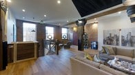 This single-story space showcases a compelling fusion of industrial and rustic design elements that together create a warm yet urban atmosphere. Exposed brick walls form a defining architectural feature throughout, lending texture and character to the interior. Original wood beams span the ceiling, reinforcing the space's historic character, while a contrasting dark-painted ceiling section adds a modern, deliberate design touch.

The flooring consists of light-toned hardwood planks that run throughout the open layout, contributing to a sense of continuity and spaciousness. Large, multi-pane windows along the exterior walls allow abundant natural light to filter in, brightening the brick surfaces and creating an inviting ambiance.

A reception or entry counter is positioned near the entrance, suggesting a welcoming, functional front-of-house area. The main open area is furnished with comfortable seating arrangements, including upholstered sofas and accent chairs with decorative pillows, creating a lounge-like environment. A prominent structural column with industrial-style steel detailing serves as a central visual focal point.

A glass-railed staircase or mezzanine opening is also visible, adding architectural depth. Overall, the space balances historic industrial character with contemporary comfort.