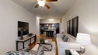 This single-floor space showcases a thoughtfully designed open-concept layout that connects the living and kitchen areas into one cohesive, inviting environment. The living room features vaulted ceilings accented by a ceiling fan, creating a sense of airiness and volume. Warm hardwood flooring flows throughout, tying the spaces together with a consistent and polished aesthetic. The living area is centered around a comfortable sectional sofa complemented by patterned accent chairs, a glass-top coffee table, and curated wall art, lending the space a contemporary yet welcoming character. A seamless transition leads into the kitchen, which is appointed with rich dark wood cabinetry, stainless steel appliances including a refrigerator and range, and a central island with bar seating — ideal for casual dining or entertaining. Pendant lighting above the island adds warmth and dimension to the kitchen, while a decorative wine rack built into the cabinetry serves as a charming design detail. Track lighting overhead ensures the kitchen is both functional and well-lit. The open sightlines between rooms enhance the sense of spaciousness, making this a practical and visually cohesive living environment well-suited for everyday comfort and social gatherings alike.
