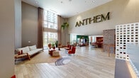 Located at Harbor Point in Stamford, CT, Anthem is a thoughtfully designed amenity space that blends contemporary style with a warm, inviting atmosphere. The grand lobby greets visitors with soaring double-height ceilings, floor-to-ceiling windows that flood the space with natural light, and wide-plank wood-look flooring that runs throughout. A bold living moss "ANTHEM" sign anchors the feature wall, while lush hanging and potted greenery adds a fresh, organic touch. The reception area features a stylishly patterned desk set against an exposed brick accent wall, a design element that recurs throughout the space to add texture and warmth. A casual lounge area near the entrance offers relaxed seating with eclectic furnishings, including woven pendant lights and a wall of built-in display niches surrounding a large flat-screen TV. The game room is a standout highlight, complete with a pool table, shuffleboard, foosball, hanging hammock chairs, a putting green, and a glowing neon "Life is Sweet" sign against a brick backdrop. Additional seating nooks and open flex areas with vibrant orange planters and railings provide versatile gathering spaces, all bathed in abundant natural light from expansive windows overlooking the Harbor Point neighborhood.